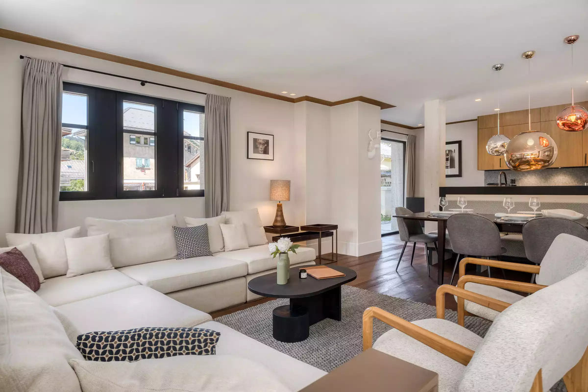 A modern living room featuring a large white sectional sofa with neutral-toned pillows, a black oval coffee table, and a gray area rug. The room has white walls with wood trim, large windows with gray curtains, and an open view to a dining area in the background.