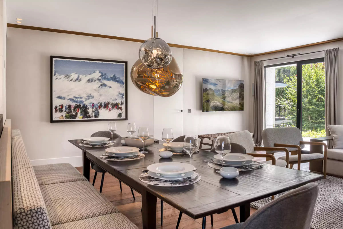 A modern dining room featuring a dark wood rectangular table set with white plates and glassware, a large textured metallic pendant light, and mountain landscape wall art.
