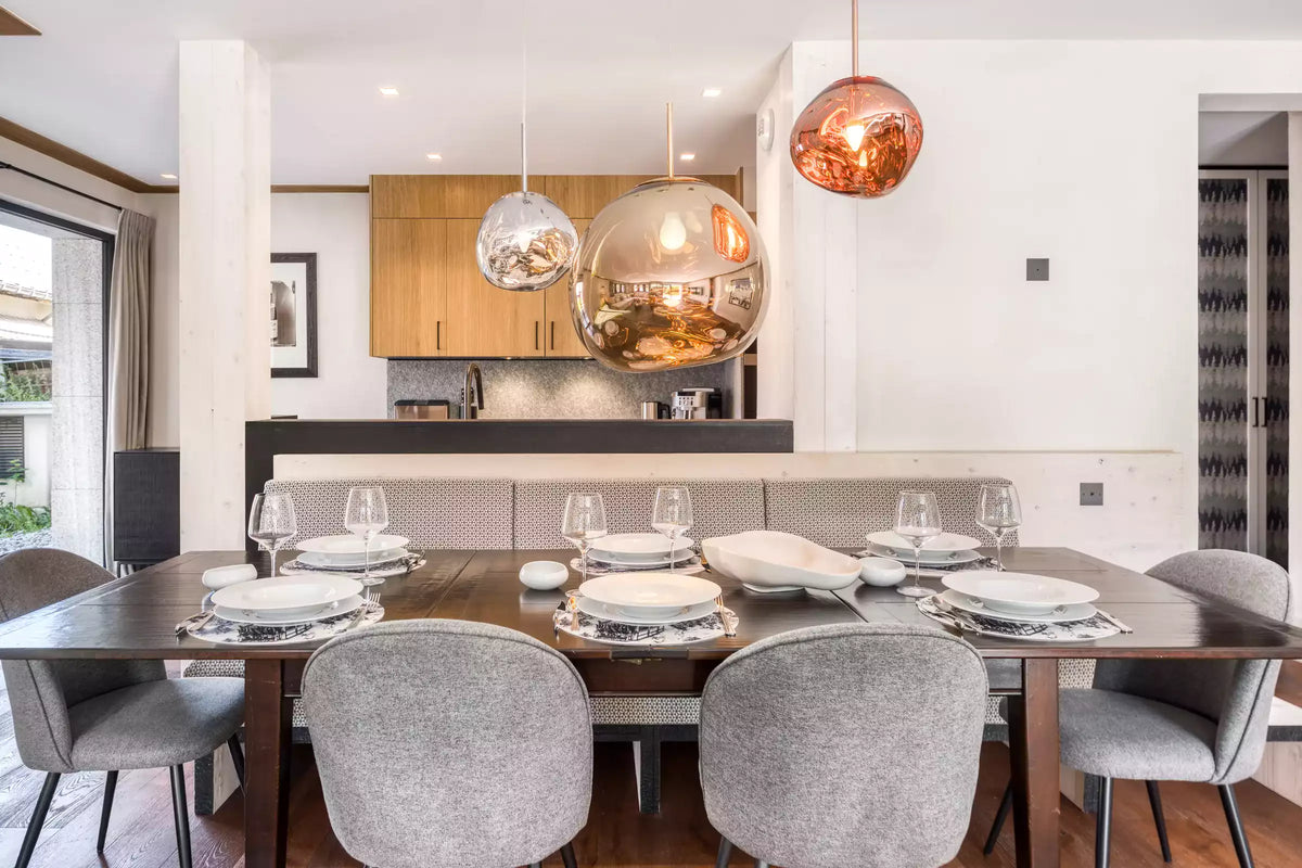 A modern dining room setup featuring a dark wood rectangular table set with white plates and wine glasses, surrounded by gray upholstered chairs. Three large, reflective spherical pendant lights hang over the table, with a kitchen area in the background.