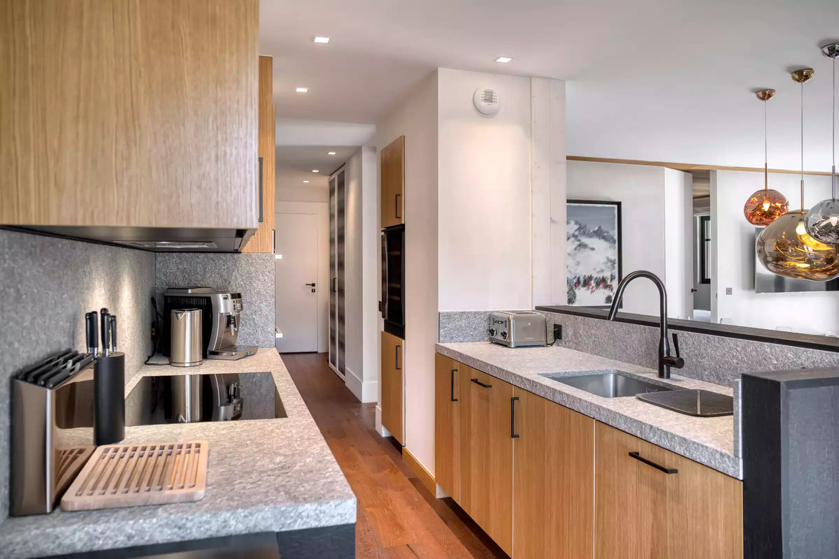 A modern kitchen interior with light wood cabinets, gray stone countertops, a built-in black cooktop, stainless steel sink and faucet, and pendant lights in the background.