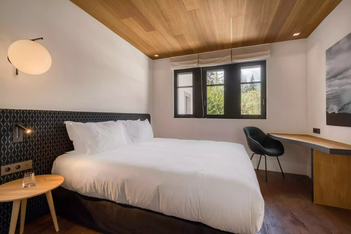 A modern bedroom interior featuring a neatly made bed with white bedding, a dark patterned headboard, a light wood ceiling, a window with black frames, a black chair, and a wooden desk against a white wall.