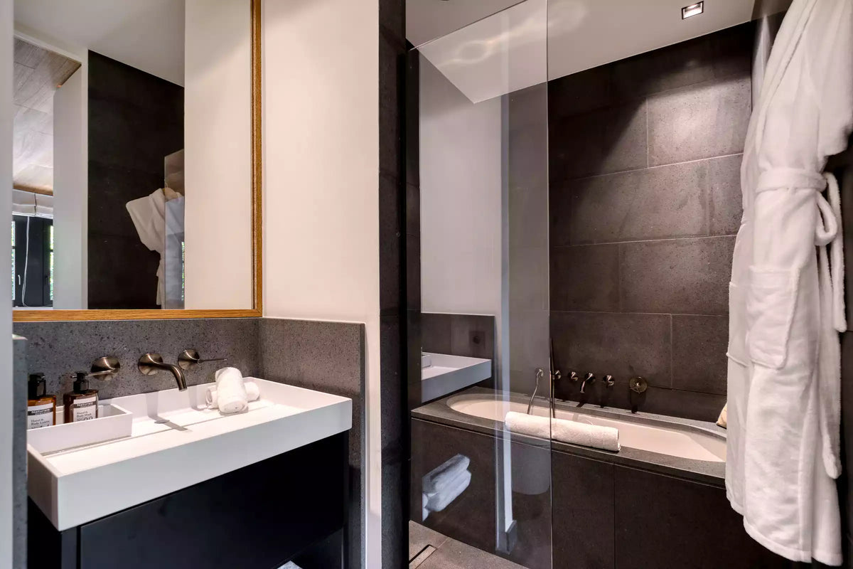 Modern bathroom interior with a white double-sink vanity, dark stone wall, large mirror, bathtub, and a white robe hanging on the right.