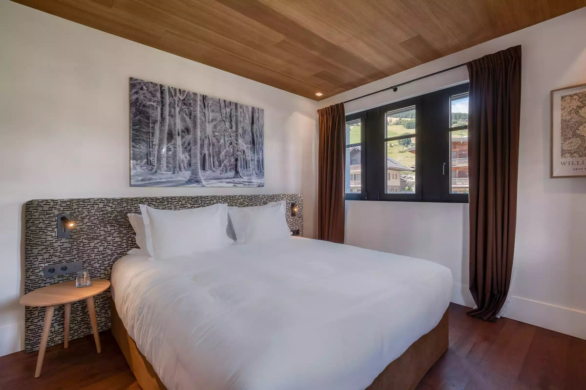 A modern bedroom featuring a large bed with crisp white bedding, a textured dark gray and beige patterned headboard, a small wooden side table, and a large window with dark brown curtains. The room has a light wood-paneled ceiling and dark wood flooring, with a framed forest print on the wall above the bed.