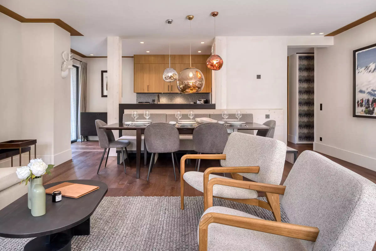 A modern living room and dining area featuring two light gray upholstered armchairs with wooden frames in the foreground, a dark round coffee table, a dining set with gray chairs, and a kitchen in the background.