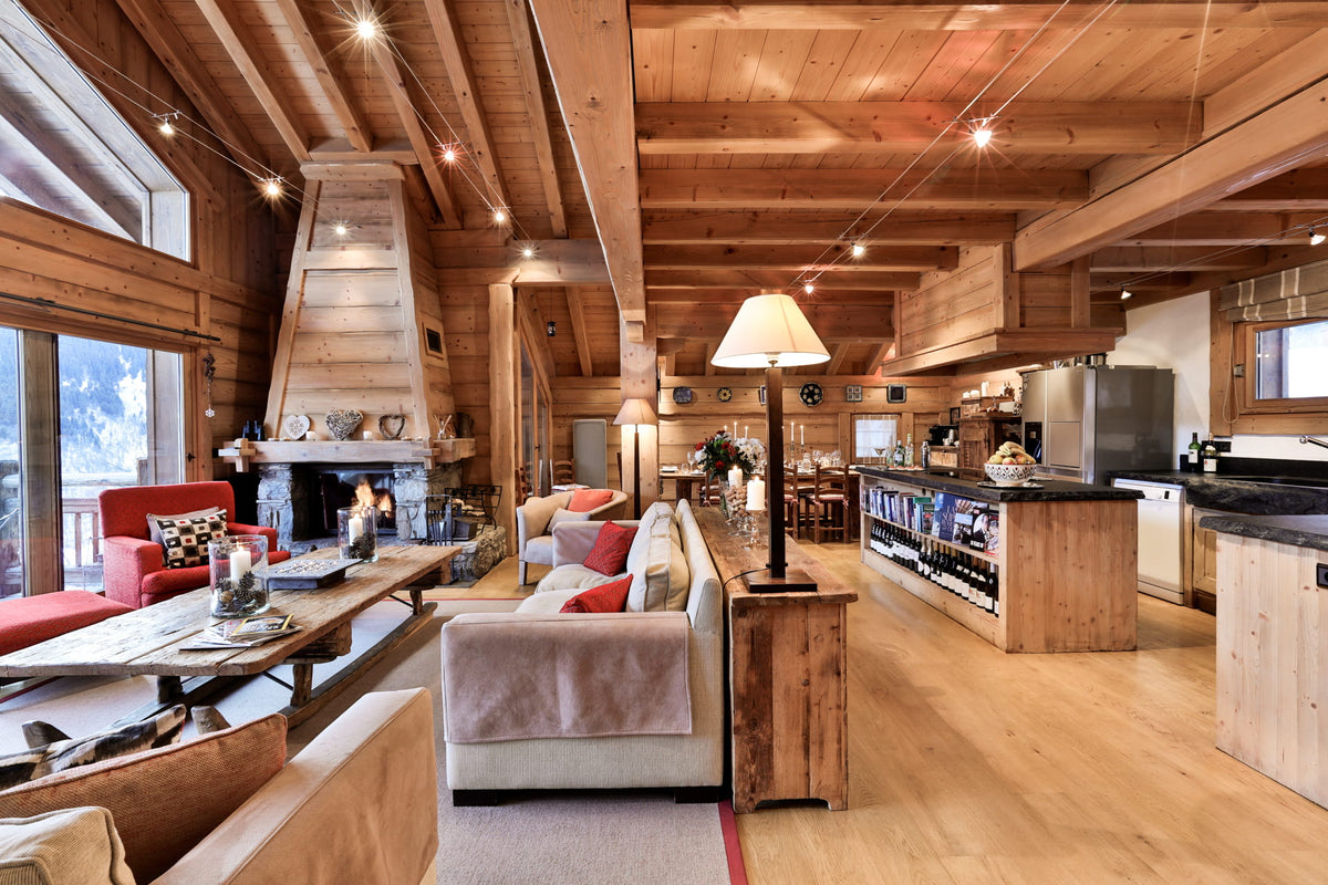 A spacious, rustic chalet-style living room with high wooden ceilings, a large stone fireplace, a light-colored sofa with red pillows, a wooden coffee table, and an open kitchen area in the background.