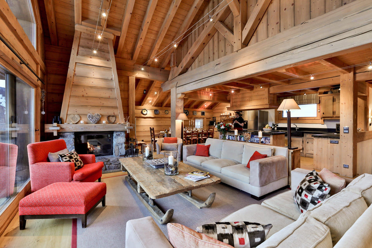 A rustic, high-ceilinged lodge-style living room with exposed wood beams, a large stone fireplace, a red armchair and ottoman, a cream sofa with red pillows, a wooden coffee table, and an open kitchen area in the background.