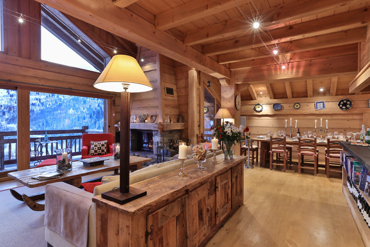 A rustic wooden chalet interior with a large wooden bar counter in the foreground, a lit table lamp, dining area with chairs and tables, fireplace, and large windows overlooking a snowy outdoor landscape.