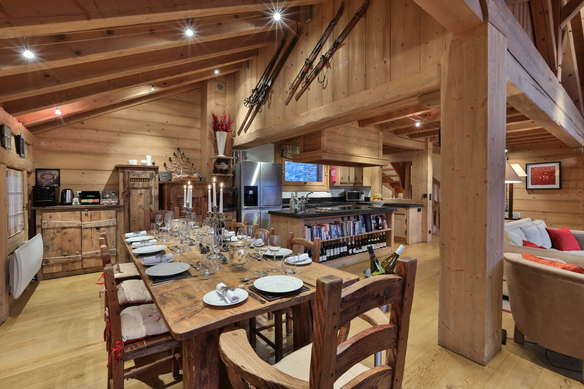 A rustic wooden dining room and kitchen interior with a long wooden table set for dining, wooden chairs, exposed wood beams, a kitchen island, and a living area in the background.