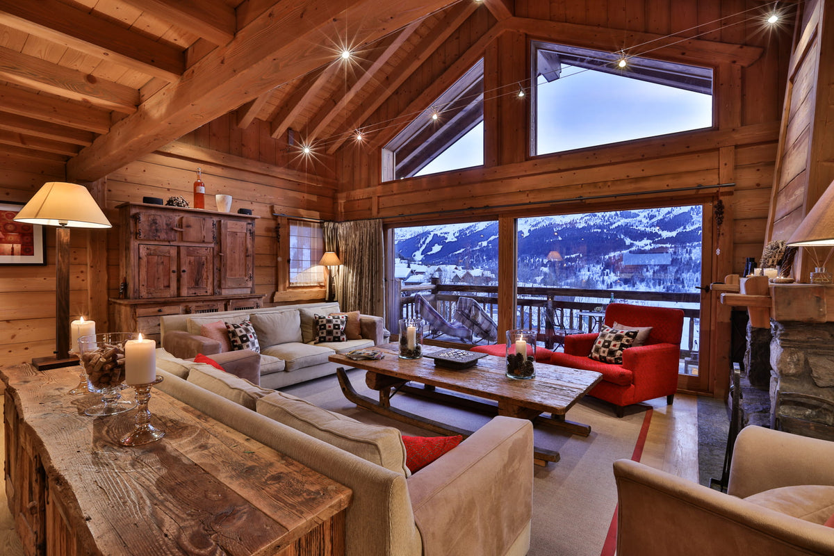 A cozy mountain chalet living room with wood-paneled walls, a large rustic coffee table, beige sofas, a red accent chair, and large windows overlooking a snowy mountain landscape.