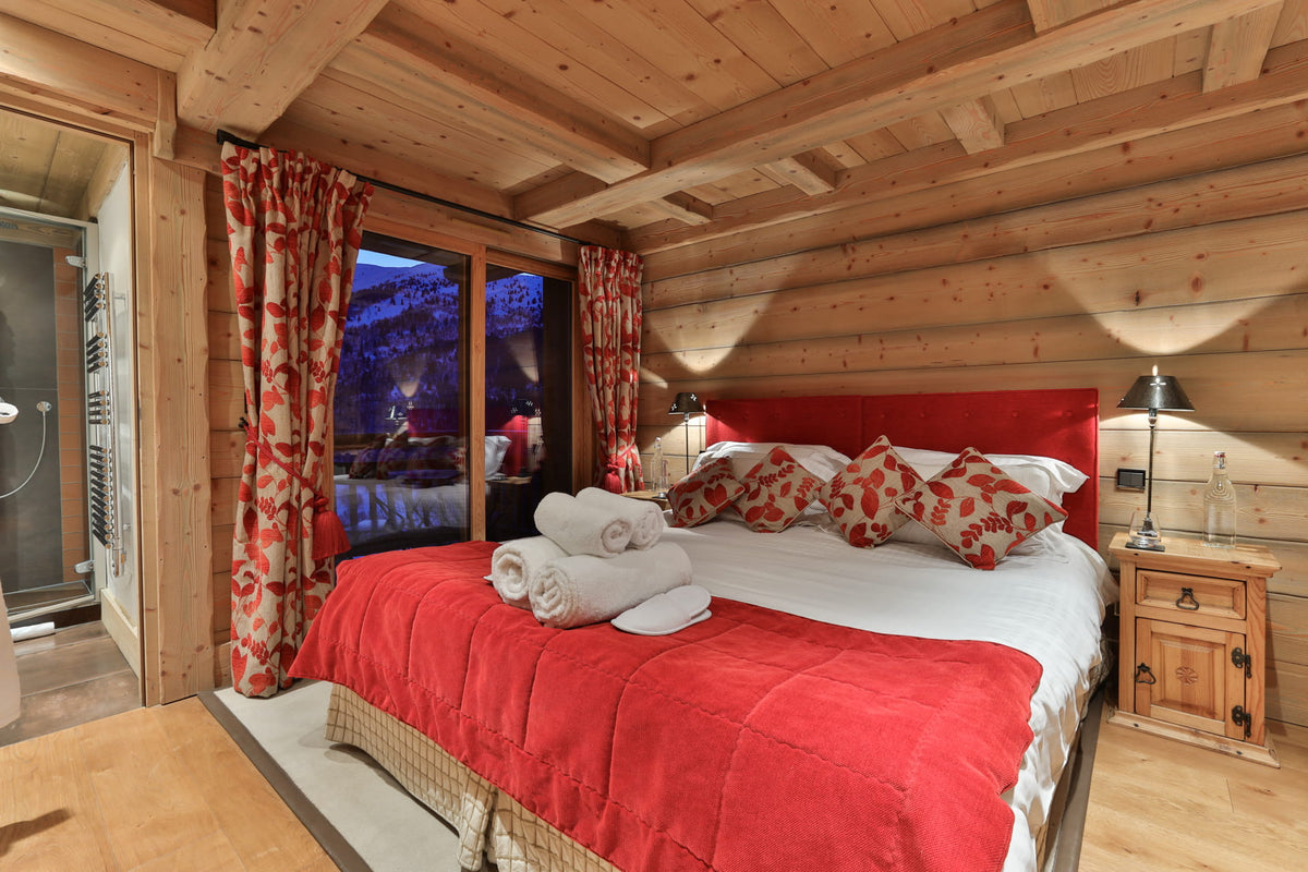 A cozy bedroom in a rustic wooden chalet, featuring a large bed with a red headboard and red quilt, white bedding, patterned red and white pillows, and matching curtains by the window.