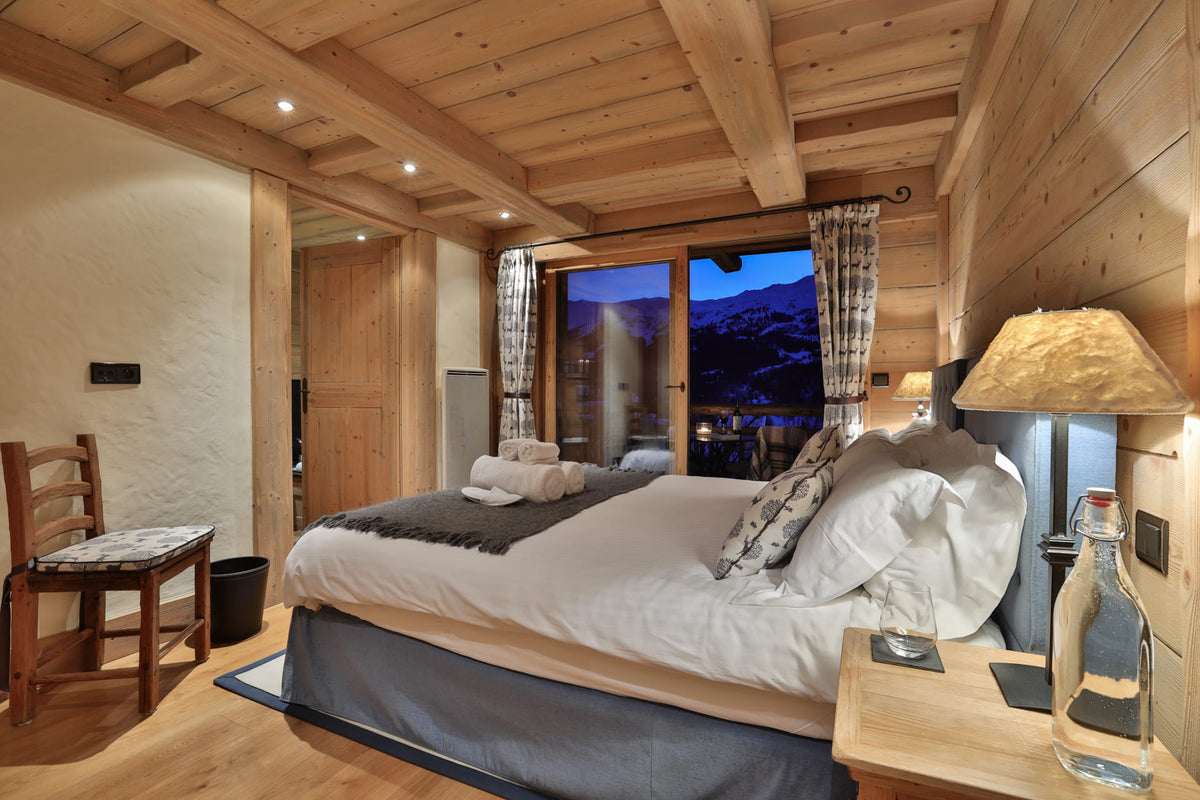 A cozy mountain chalet bedroom featuring a large bed with white linens and gray bedding, set against wood-paneled walls and ceiling. A window reveals a scenic mountain view at dusk, and a bedside table with a lamp and water bottle is visible.