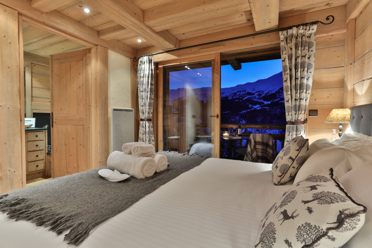 A cozy mountain chalet bedroom featuring a large bed with white linens and patterned pillows, a gray throw blanket, and a large glass window overlooking a snowy mountain landscape at dusk.