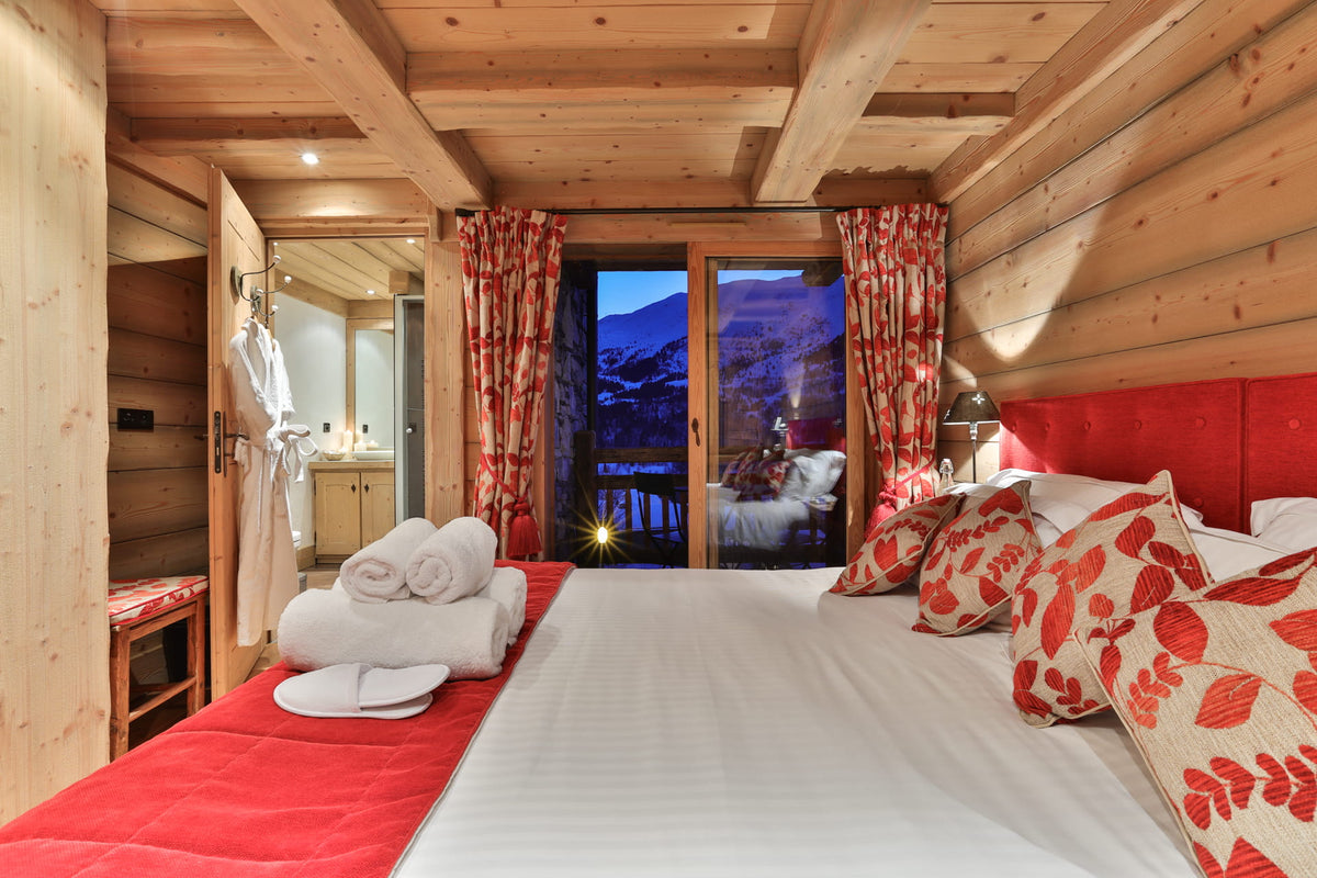 A cozy alpine-style bedroom with a large bed dressed in white linens and red-and-cream patterned pillows, set in a wood-paneled room with red curtains and a mountain view outside.