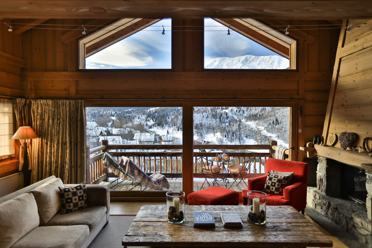 A cozy mountain chalet living room with a large rustic wooden table in the foreground, a beige sofa on the left, and a red armchair on the right, all overlooking a snowy mountain landscape from large arched windows.