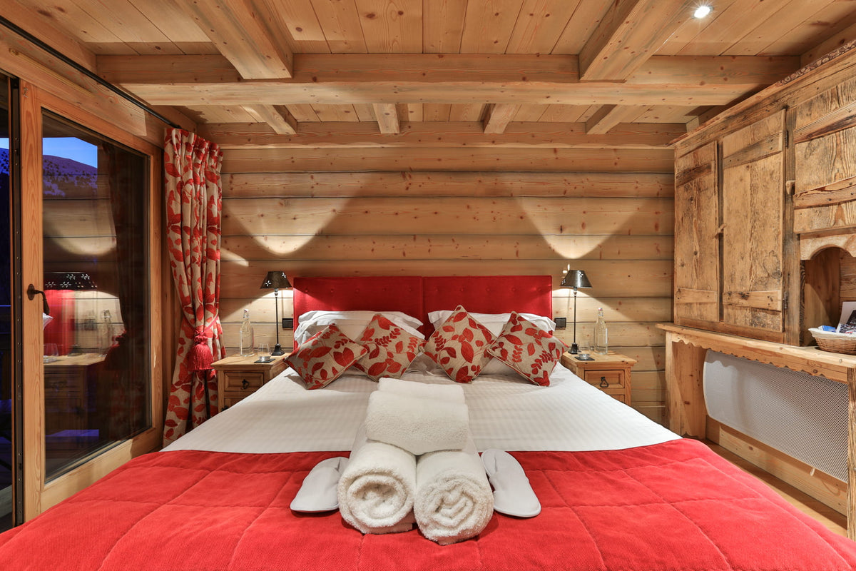 A cozy bedroom interior with a large bed dressed in white bedding and a red quilted cover, set against rustic wood-paneled walls and ceiling. Red-and-white patterned pillows, matching curtains, bedside tables with lamps, and a wooden cabinet are visible.