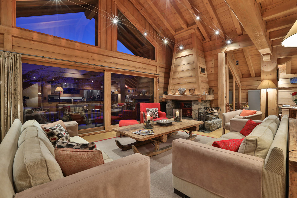 A cozy, rustic lodge-style living room with high wood-beam ceilings, large windows looking out onto a twilight sky, a stone fireplace, and multiple upholstered sofas with red and patterned pillows arranged around a wooden coffee table.