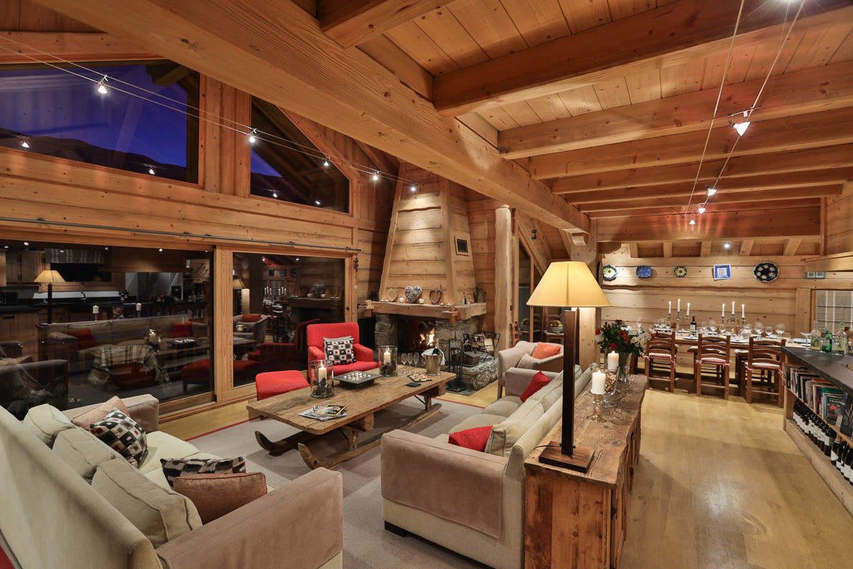 A spacious, rustic mountain chalet living room with high wooden ceilings, a large fireplace, and multiple upholstered sofas in beige and red. A wooden dining table and chairs are visible in the background, along with string lights and warm ambient lighting throughout the space.