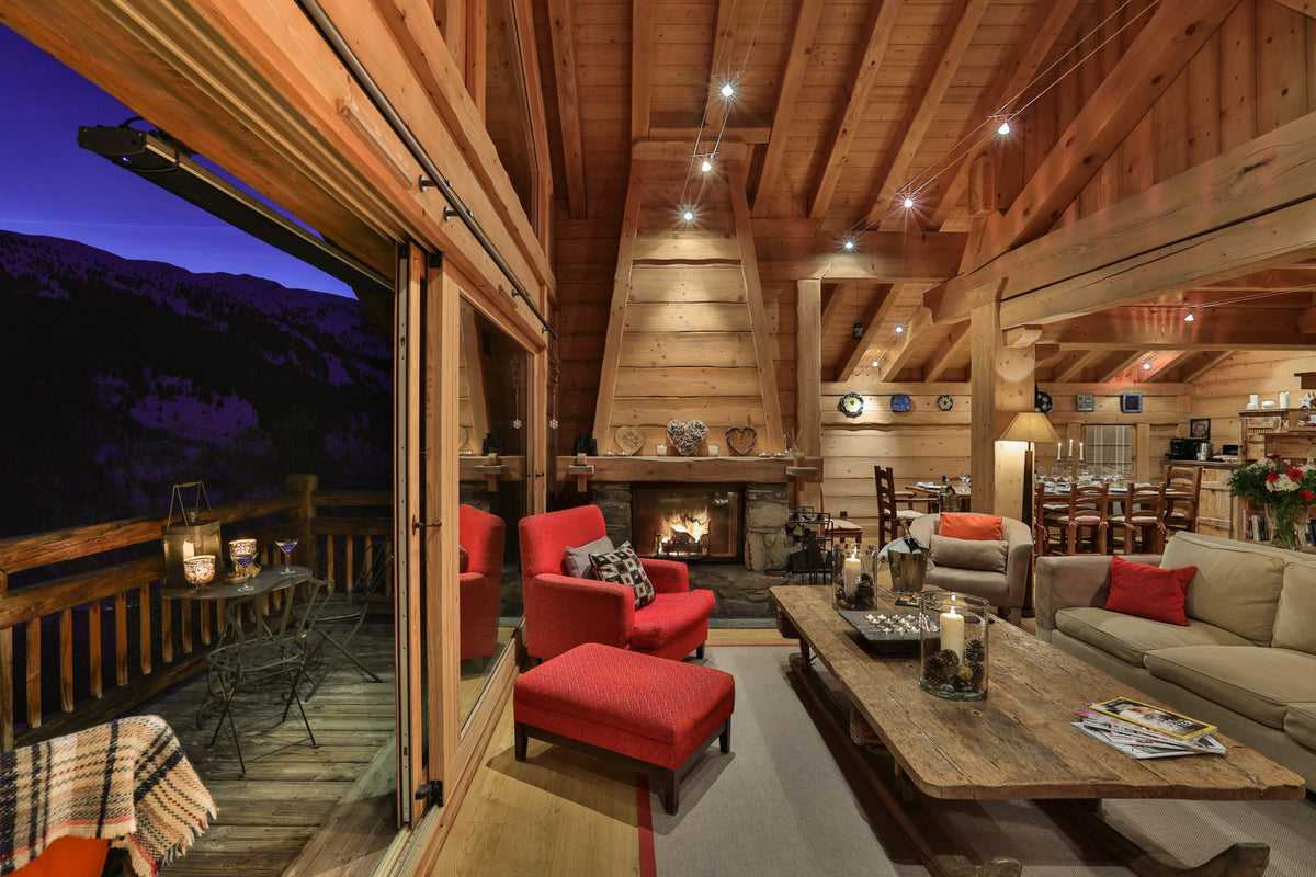 A cozy mountain chalet living room with warm wood walls and ceiling, featuring a fireplace, a beige sofa with red pillows, two red armchairs with matching red ottomans, and a rustic wooden coffee table. Large sliding glass doors open to a mountain view at dusk.