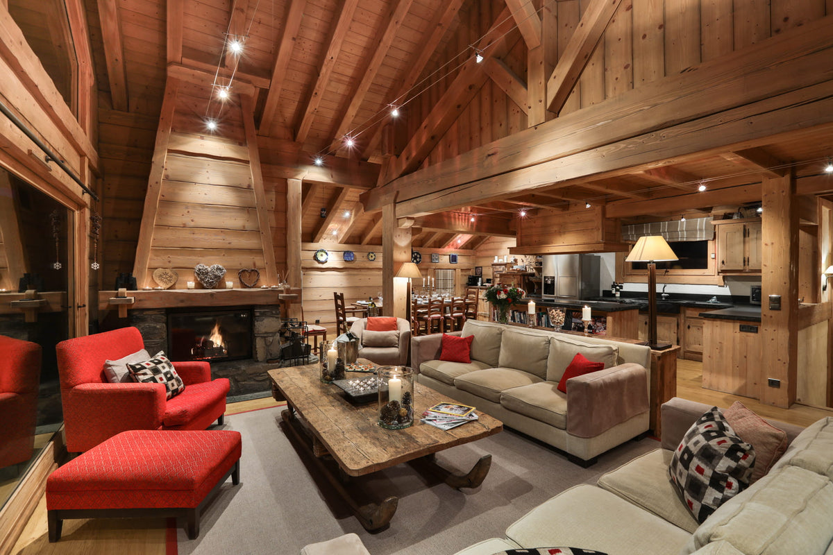 A rustic, high-ceilinged chalet-style living room with exposed wood beams, a fireplace, a wooden coffee table, a beige sofa with red accent pillows, and a red armchair with matching ottoman. A kitchen and bar area are visible in the background.