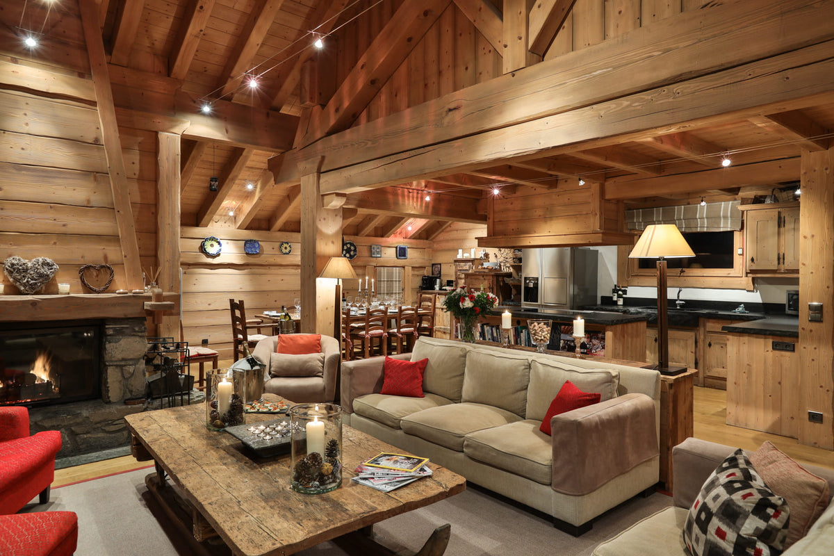 A rustic log cabin living room with a beige sofa, red accent pillows, a wooden coffee table, a fireplace, and an open kitchen area in the background.