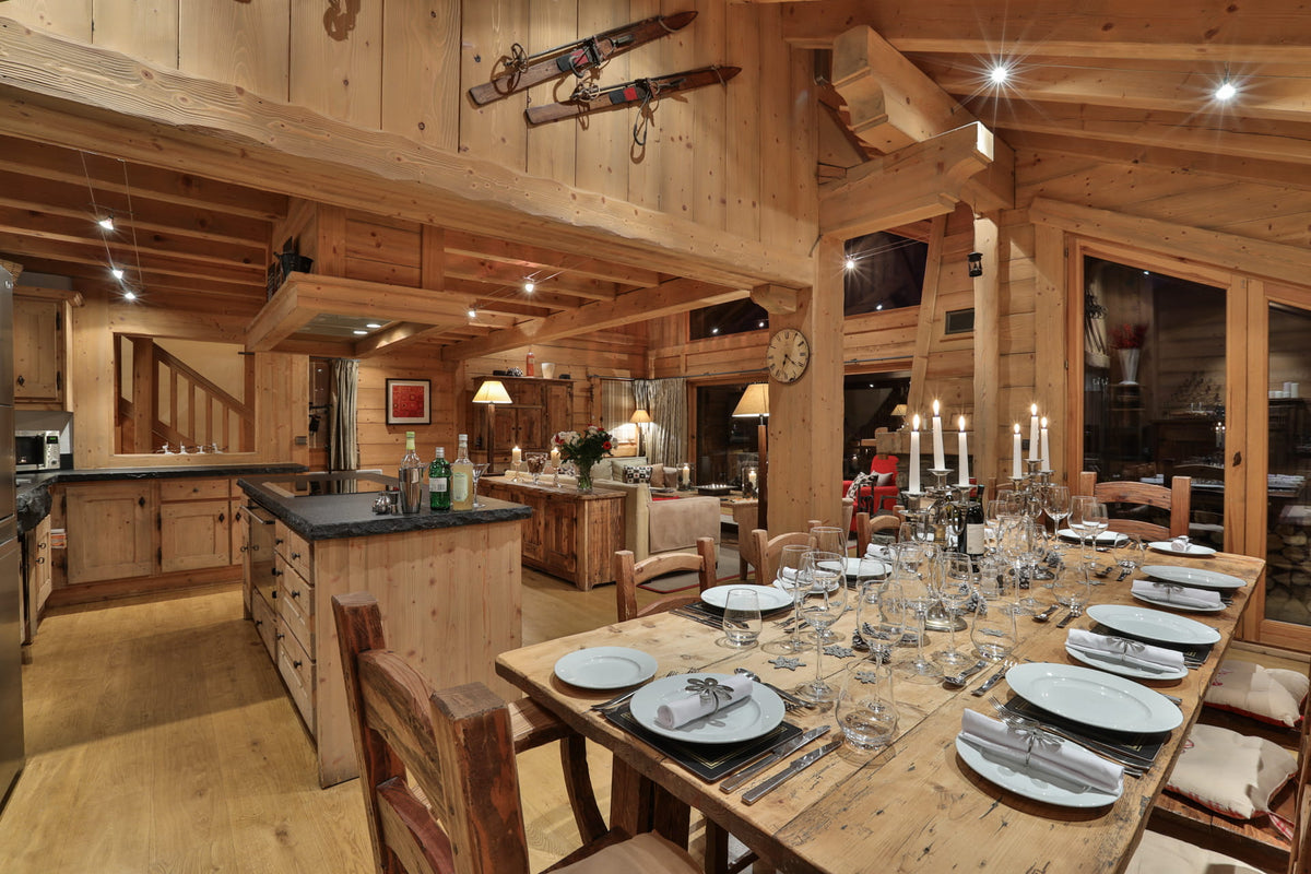 A rustic alpine chalet interior featuring a large wooden dining table set with white plates, glassware, and candles, surrounded by wooden chairs. In the background, a matching wood kitchen island and cabinetry are visible under warm ceiling lights.
