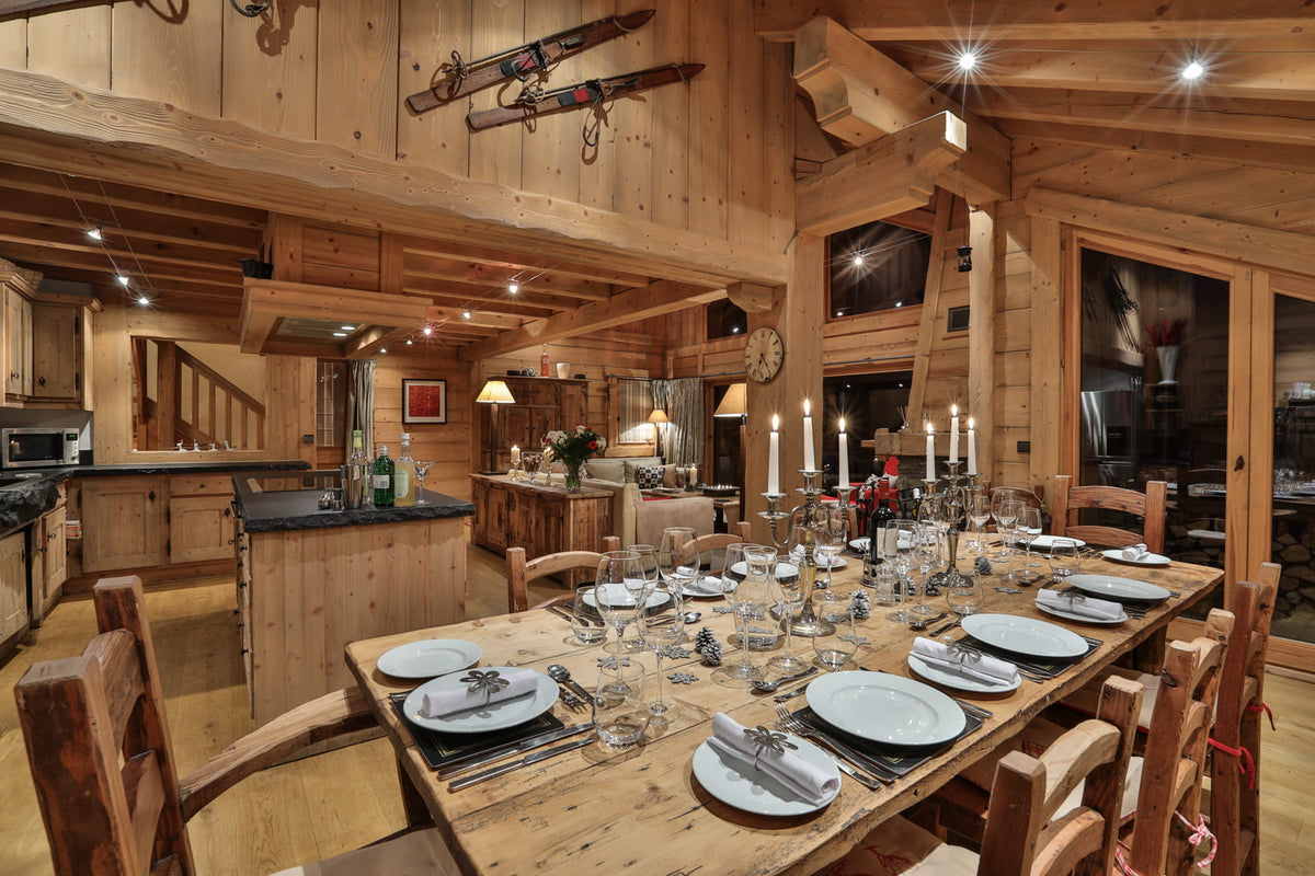 A rustic wooden dining room and kitchen interior with a long wooden dining table set for multiple guests, surrounded by matching wooden chairs. The space features exposed wood beams, wood-paneled walls, and a kitchen area in the background with cabinetry and countertops.