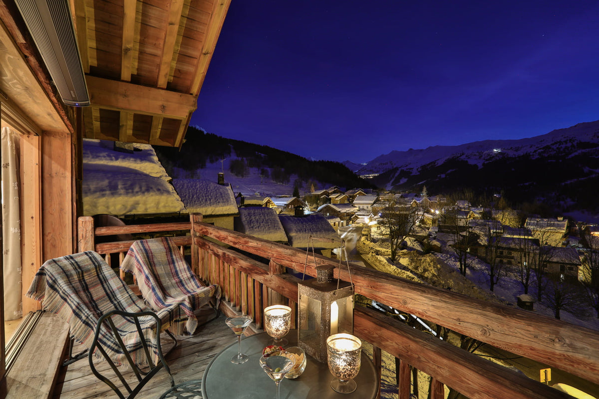 A cozy wooden balcony at night overlooking a snowy mountain village under a deep blue twilight sky. Two plaid lounge chairs sit on the deck beside a small table with lit candles and glasses, with mountains and chalets visible in the background.