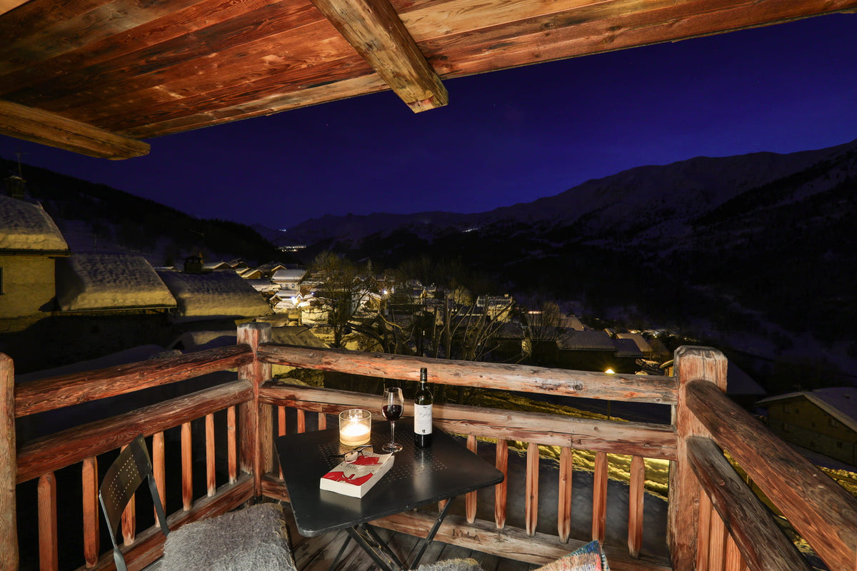 A cozy wooden balcony overlooking a mountain village at night, with a small table holding a bottle, two wine glasses, and a lit candle, set against a deep blue mountain landscape.