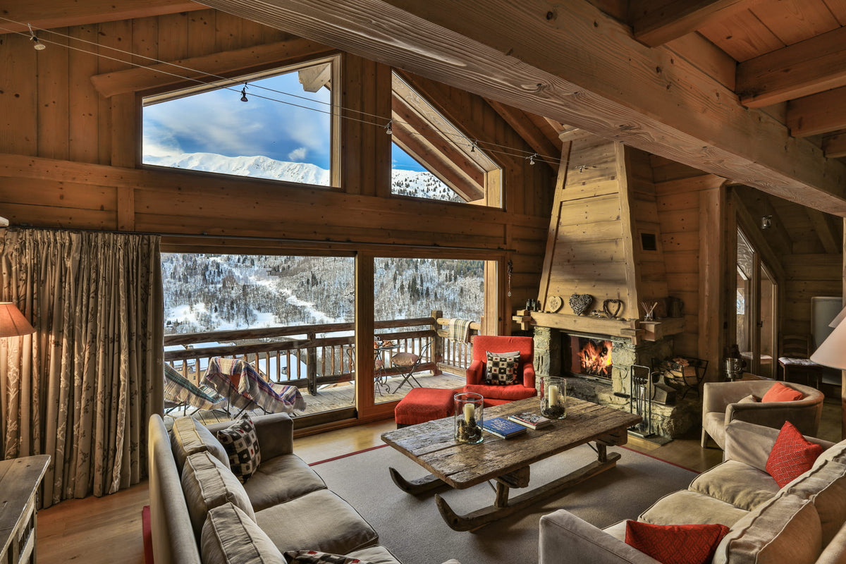 A cozy mountain chalet living room with wood-paneled walls, a large stone fireplace, tall windows overlooking a snowy mountain landscape, and upholstered seating including a gray sofa, armchair, and red accent chair.