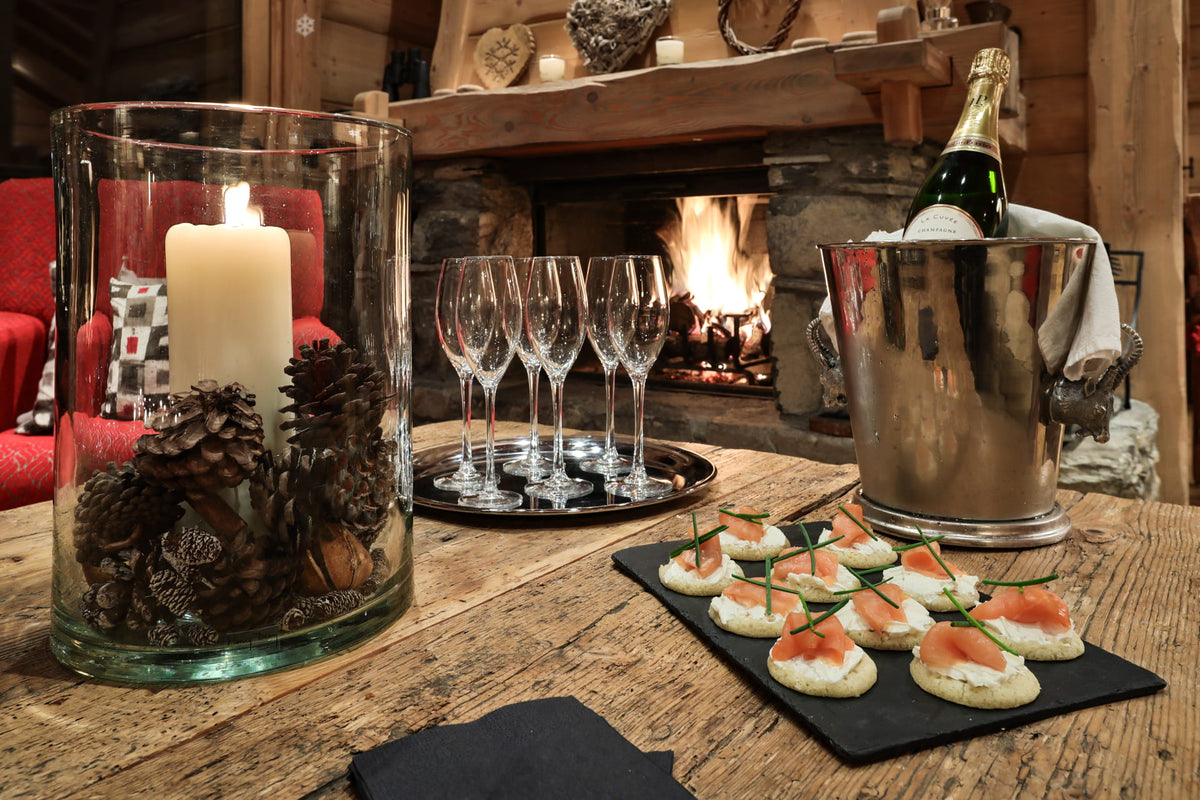 A cozy indoor table setting by a fireplace with a large glass candle holder containing pinecones and a lit white pillar candle, a metal wine bucket with champagne bottle, four stemmed wine glasses on a tray, and canapes on a black slate board.