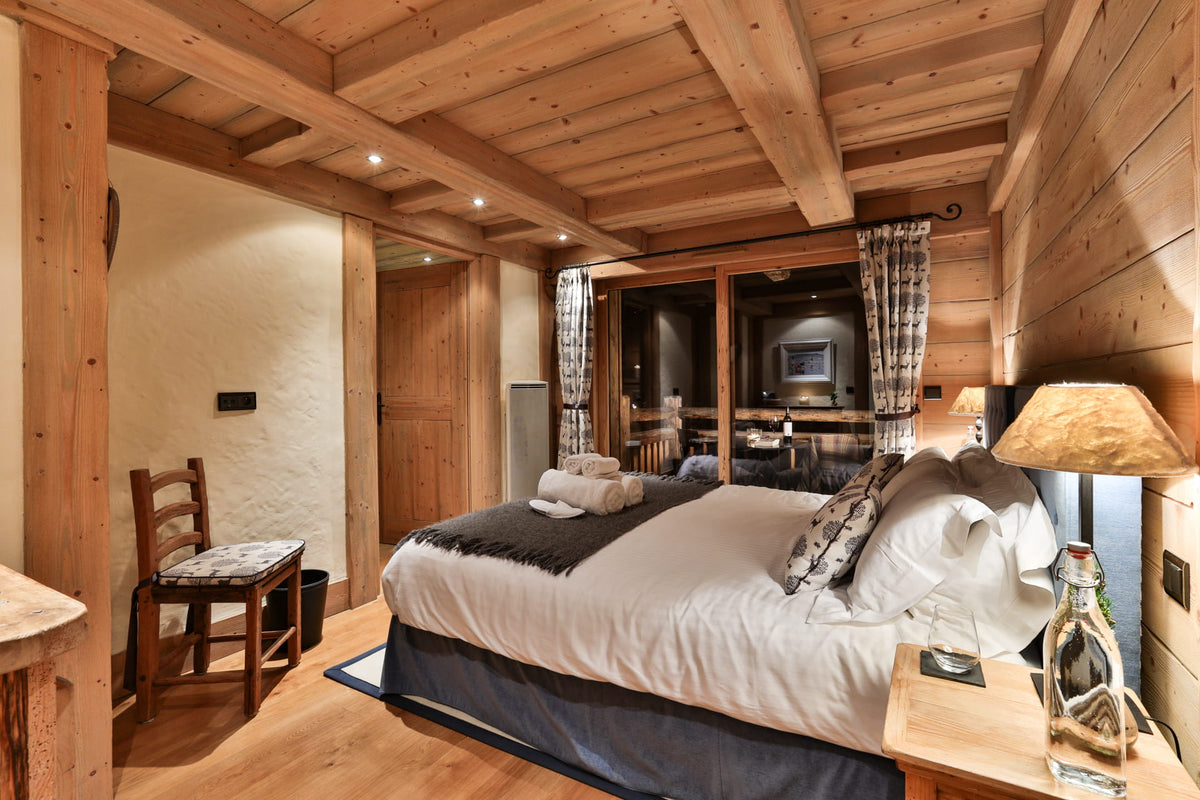 A cozy, rustic bedroom with a large bed dressed in white bedding and dark gray accents, set in a wooden chalet with exposed wood ceiling beams, a wooden chair, and a bedside table with a lamp.