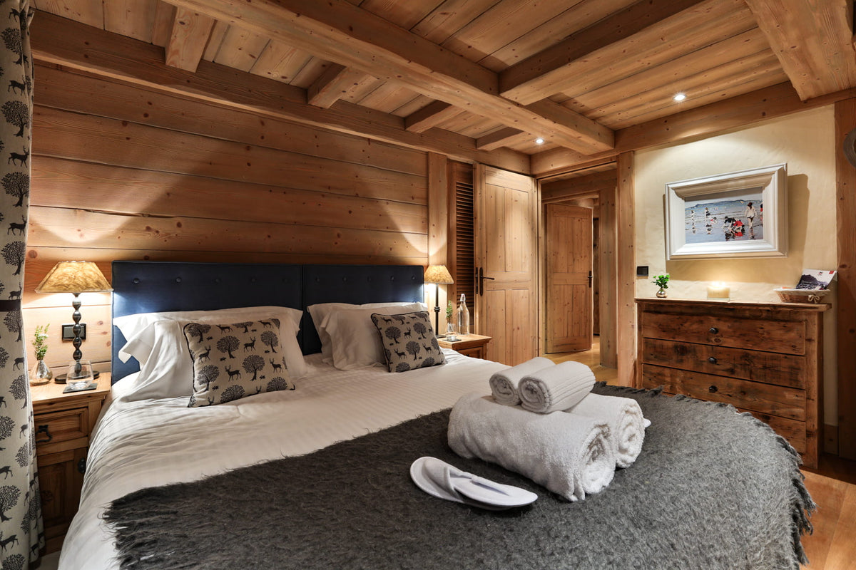 A cozy, rustic bedroom with a large bed dressed in white linens, patterned pillows, a dark blue headboard, and a gray throw blanket. The room features warm wood-paneled walls and ceiling, wooden nightstands with lit lamps, a wooden dresser, and rolled towels on the bed.