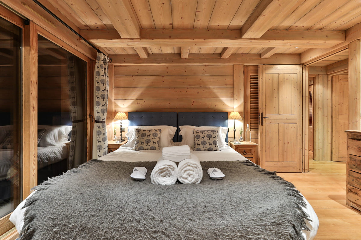 A cozy bedroom with a large bed dressed in white linens and a gray textured blanket, set in a rustic wooden chalet with a blue headboard, bedside lamps, and a glass sliding door on the left.