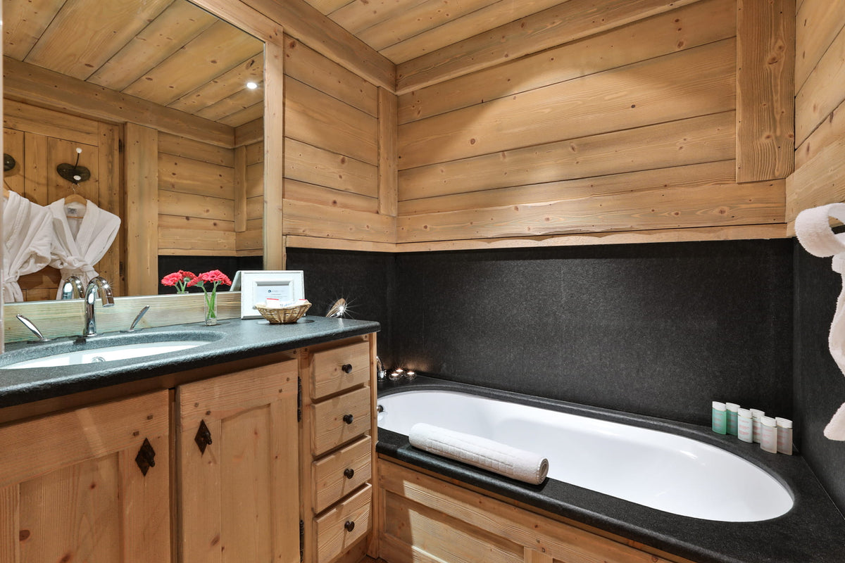 A luxury spa-style bathroom featuring a rustic light wood cabinetry design, a black stone countertop, a white oval bathtub, and a large wall-mounted mirror.