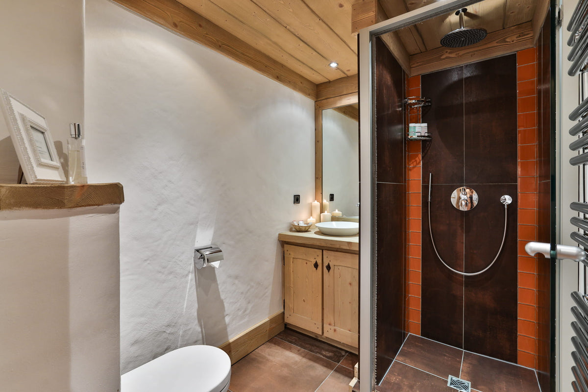 A rustic modern bathroom featuring a walk-in shower with dark wood panels and terracotta brick walls, a white toilet, a wooden vanity with a white sink, and textured white plaster walls.
