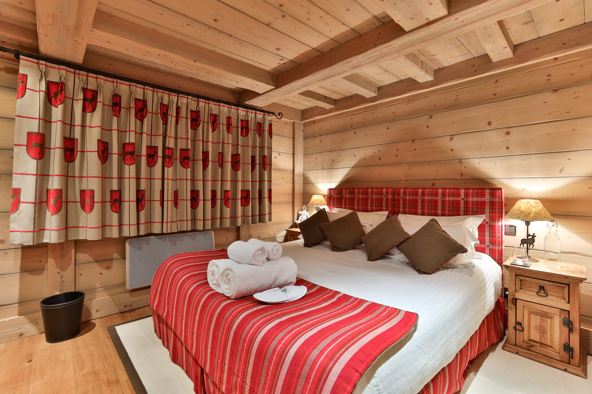 A cozy bedroom in a rustic wood-paneled chalet with a double bed dressed in red and white striped bedding, brown pillows, and a red plaid headboard. Matching patterned curtains hang on the window, and wooden nightstands with lamps sit on either side.