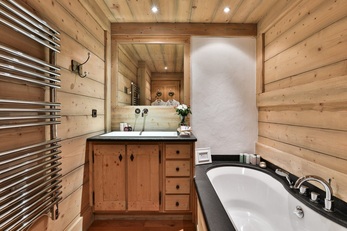 A luxury bathroom interior featuring light natural wood paneling on the walls and ceiling, a wooden vanity with a white sink, a large mirror, a heated towel rail, and a white bathtub with a black countertop.