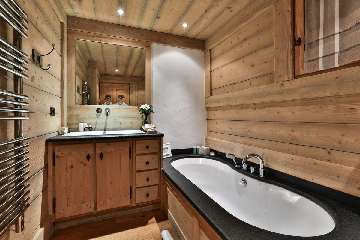 A luxury alpine-style bathroom featuring a double vanity with a white basin, a large white double sink, a white bathtub with a black countertop, and warm wood-paneled walls and ceiling.