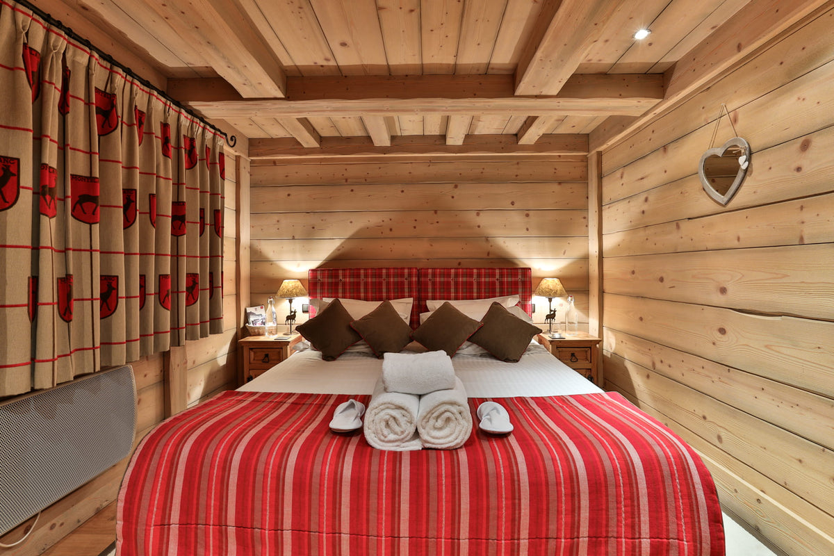 A cozy bedroom with light wood paneled walls and ceiling, featuring a bed dressed in a red and white striped blanket, brown pillows, and matching patterned curtains.