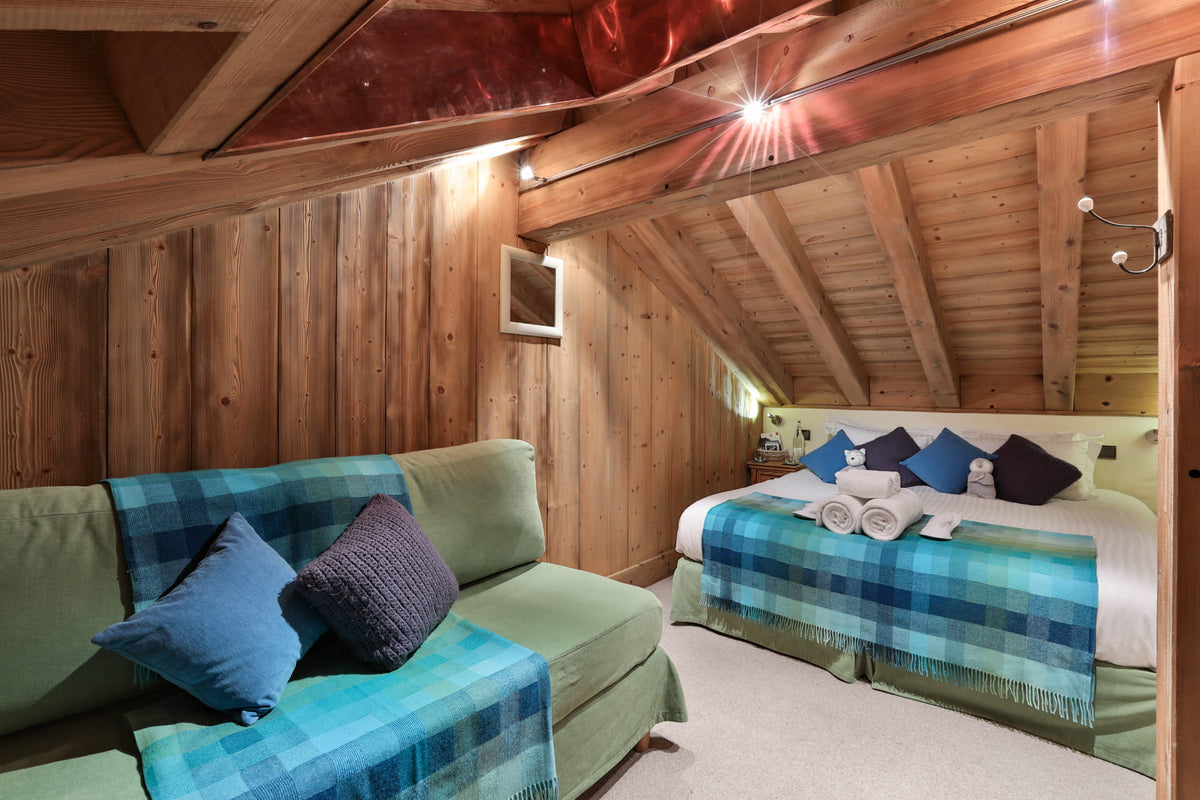 A cozy attic bedroom with wood-paneled walls and slanted ceiling, featuring a green sofa with blue plaid throw pillows and blanket, and a bed with matching blue plaid bedding and white towels.