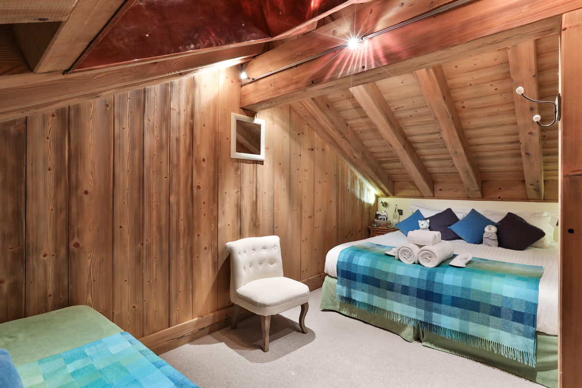 A cozy attic bedroom with wood-paneled walls and slanted ceiling beams, featuring a bed with blue and green plaid bedding, a white armchair, and warm overhead lighting.