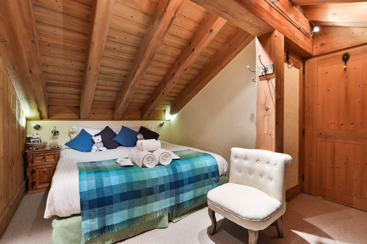 A cozy bedroom with a wooden slatted ceiling and walls, featuring a bed with blue and green plaid bedding, a white tufted armchair, and warm ambient lighting.