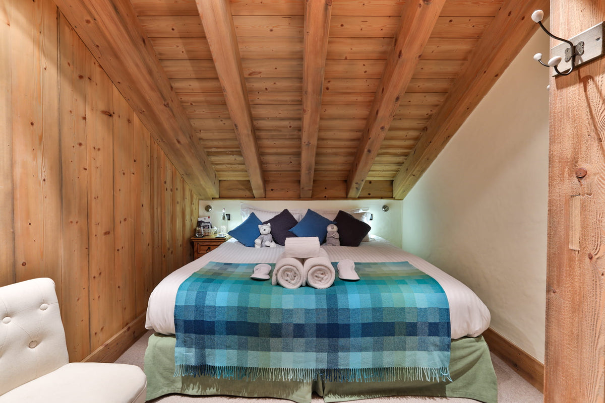 A cozy bedroom with a bed dressed in white linens, blue and navy pillows, and a blue plaid throw blanket. The room features wood-paneled walls and a wooden slanted ceiling with exposed beams.