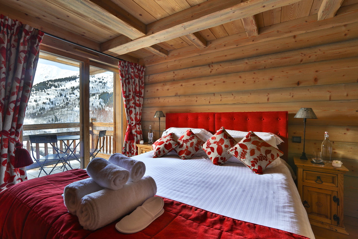 A cozy ski chalet bedroom with a large bed dressed in white and red bedding, red tufted headboard, matching pillows, rolled towels, and slippers. The room has wood-paneled walls and ceiling, a window overlooking a snowy mountain view, and bedside tables with lamps.
