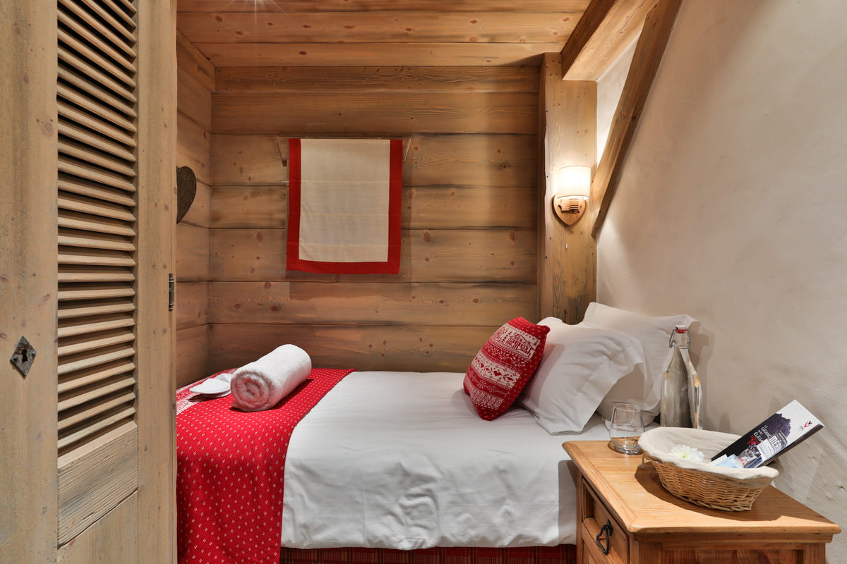 A cozy bedroom interior with a neatly made bed, white bedding, red accents, wooden walls, and a bedside table holding a basket, bottle, and magazine.