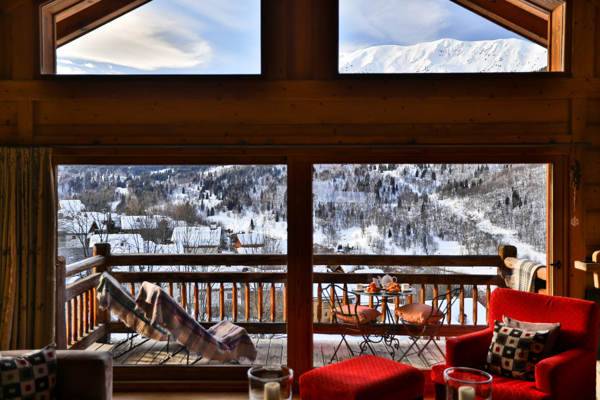 A cozy interior looking out onto a snowy mountain valley from a large wooden window. The room has warm wood framing, a red armchair, and a matching red ottoman, with outdoor snow-covered hills and trees visible beyond.