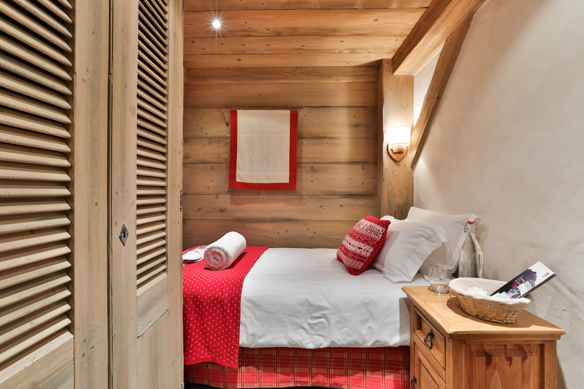 A cozy bedroom interior with a neatly made bed, white bedding, red patterned accents, wooden walls and ceiling, a wooden nightstand, and a slatted wooden wardrobe door on the left.