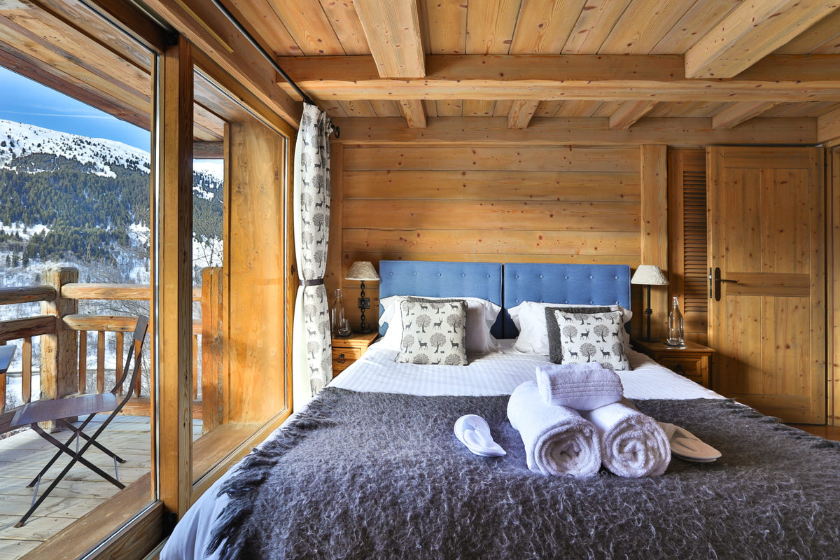 A cozy mountain chalet bedroom with a large bed dressed in gray and white linens, a blue upholstered headboard, wooden walls and ceiling, and a balcony overlooking snowy mountains.