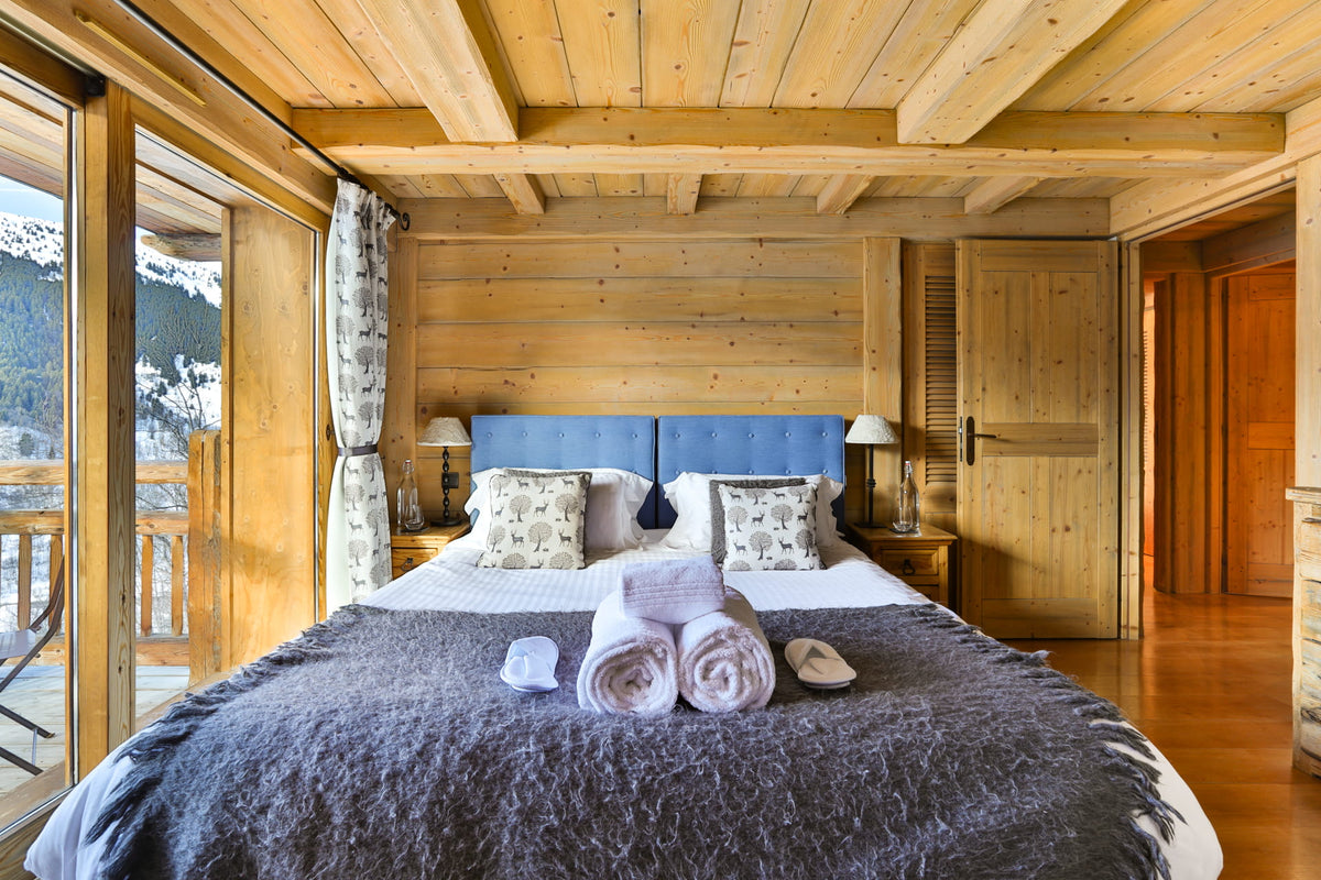 A cozy mountain chalet bedroom featuring a large bed with a blue headboard, white and gray bedding, and rolled towels on the floor. The room has light wood walls, ceiling, and flooring, with large windows showing a snowy mountain landscape outside.