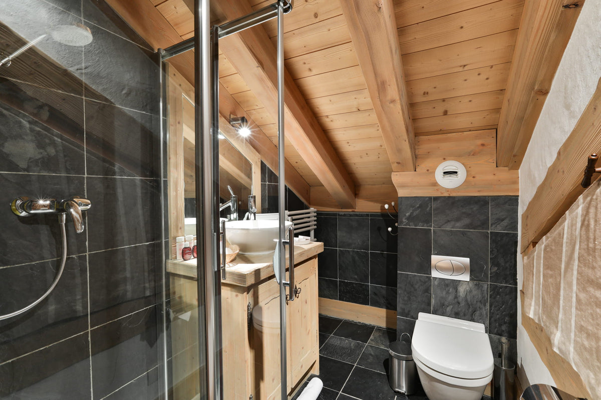 A modern bathroom with a walk-in shower, white toilet, wooden ceiling beams, and dark gray tiled walls.
